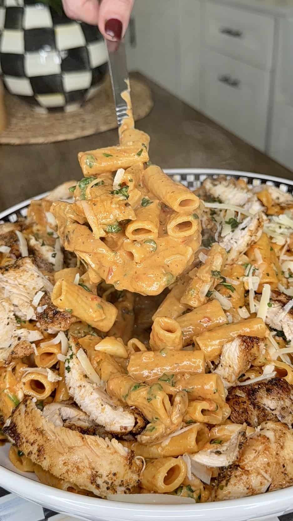 A fork lifts a serving of creamy rigatoni pasta with herbs from a bowl topped with grilled chicken strips and shredded cheese, set on a kitchen counter.