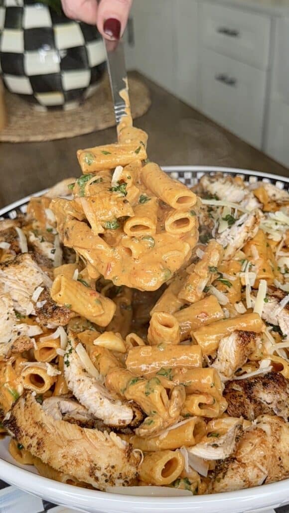 A fork lifts creamy rigatoni pasta mixed with herbs above a plate topped with sliced grilled chicken and garnished with grated cheese, set on a kitchen counter.