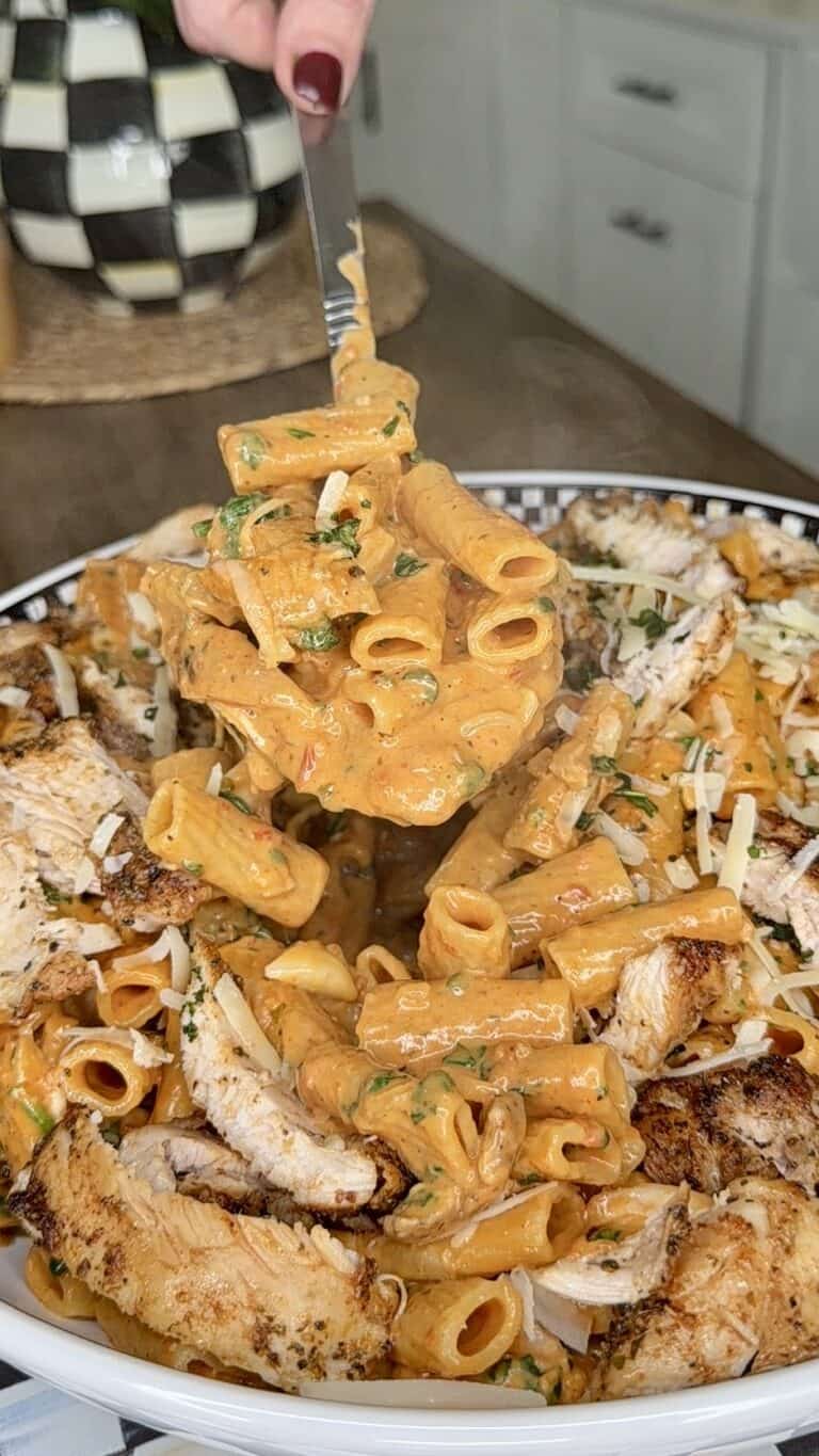 A fork lifts creamy rigatoni pasta mixed with herbs above a plate topped with sliced grilled chicken and garnished with grated cheese, set on a kitchen counter.