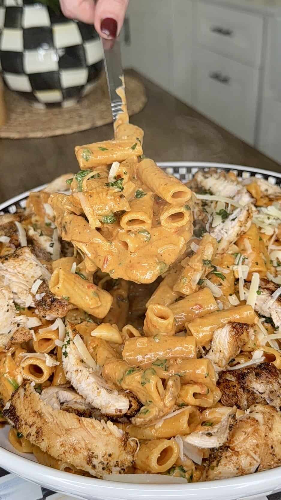 A fork lifts creamy rigatoni pasta mixed with herbs above a plate topped with sliced grilled chicken and garnished with grated cheese, set on a kitchen counter.