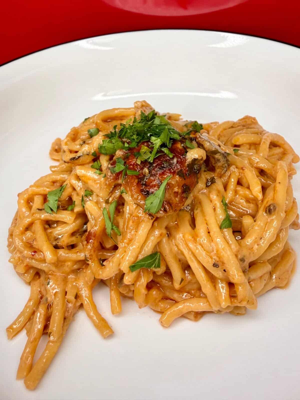 A close-up of creamy marry me baked spaghetti topped with herbs, served on a white plate with a red background. The pasta appears rich in sauce, with green garnish sprinkled on top.