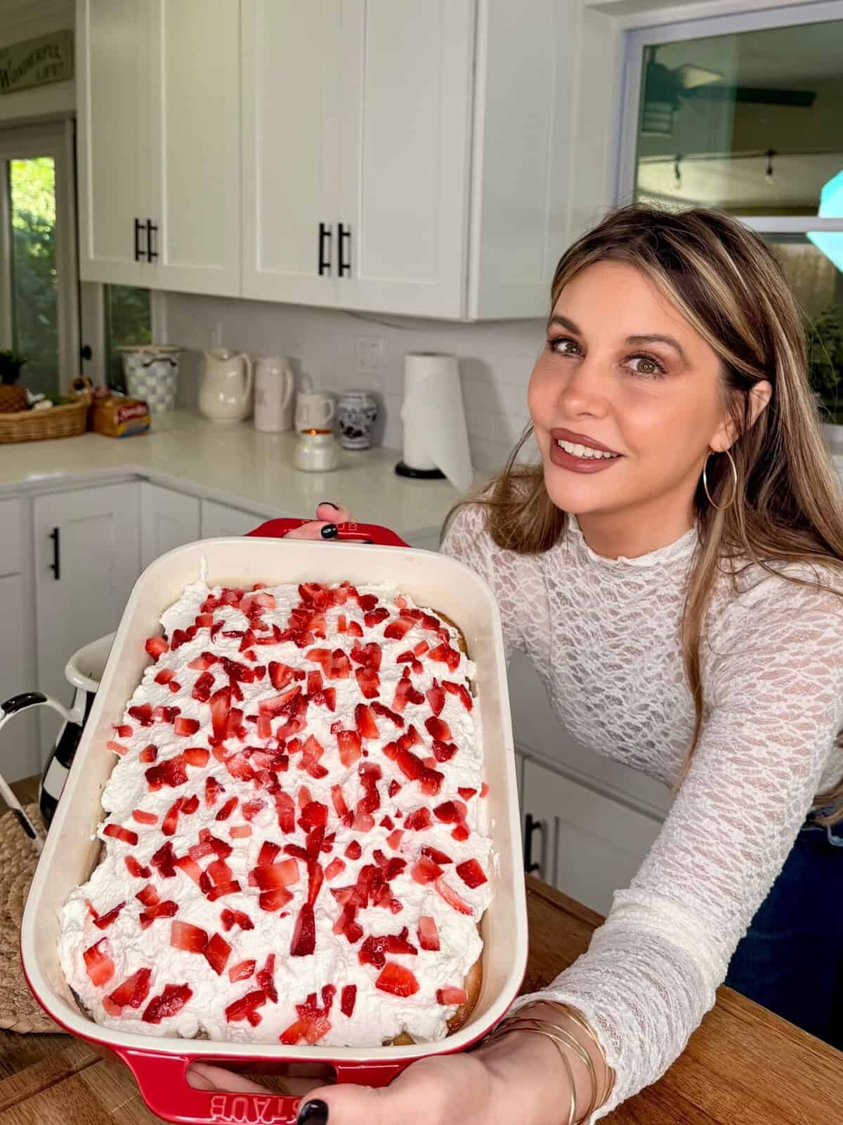 A woman in a white lace top smiles as she holds a rectangular dish filled with Easy Strawberry Tres Leches Poke Cake, topped with whipped cream and chopped strawberries, in a bright kitchen.