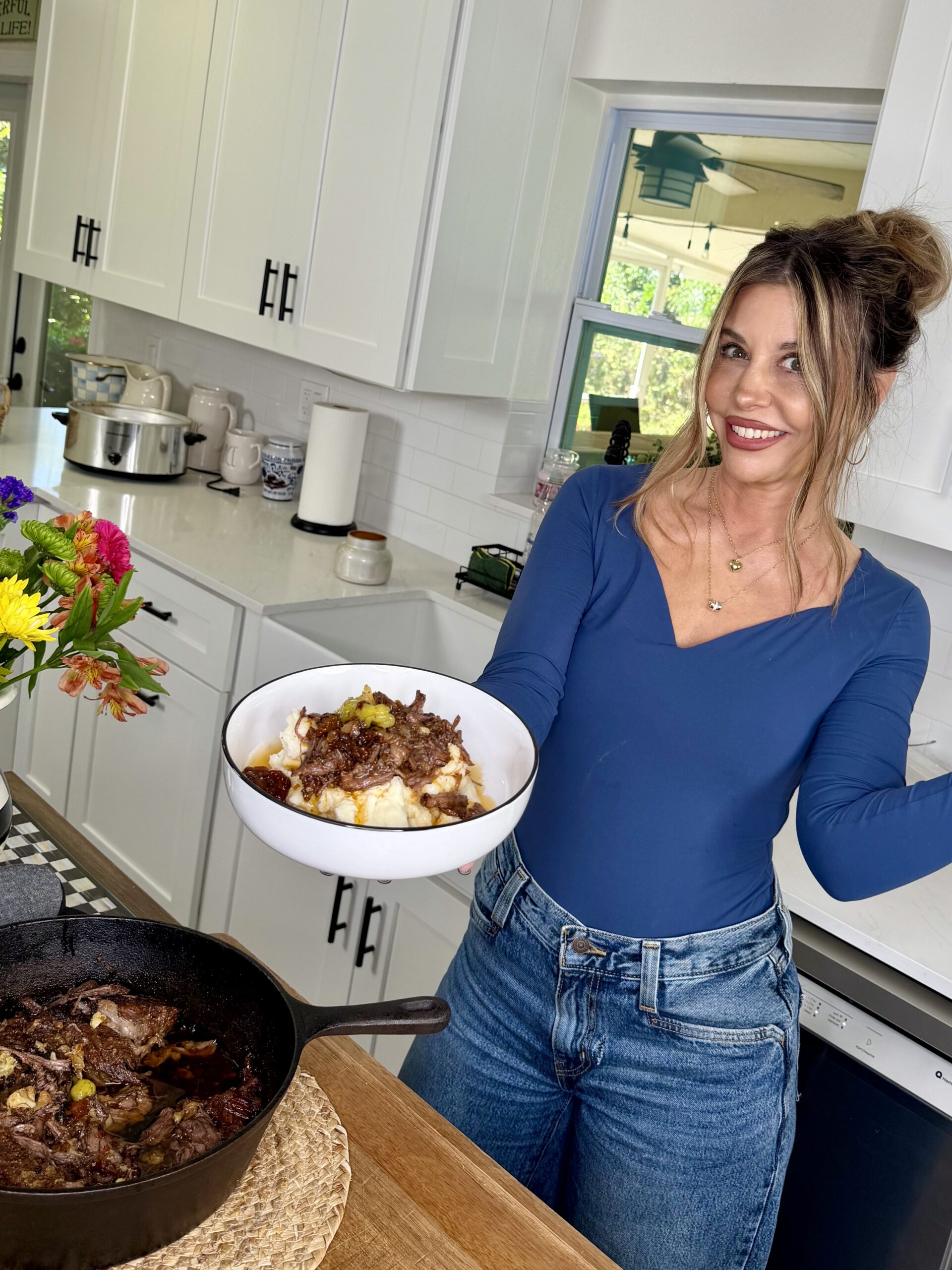 A woman in a blue shirt and jeans smiles in a bright kitchen while holding a bowl of food; a skillet with cooked meat sits on the counter nearby, and flowers decorate the kitchen.