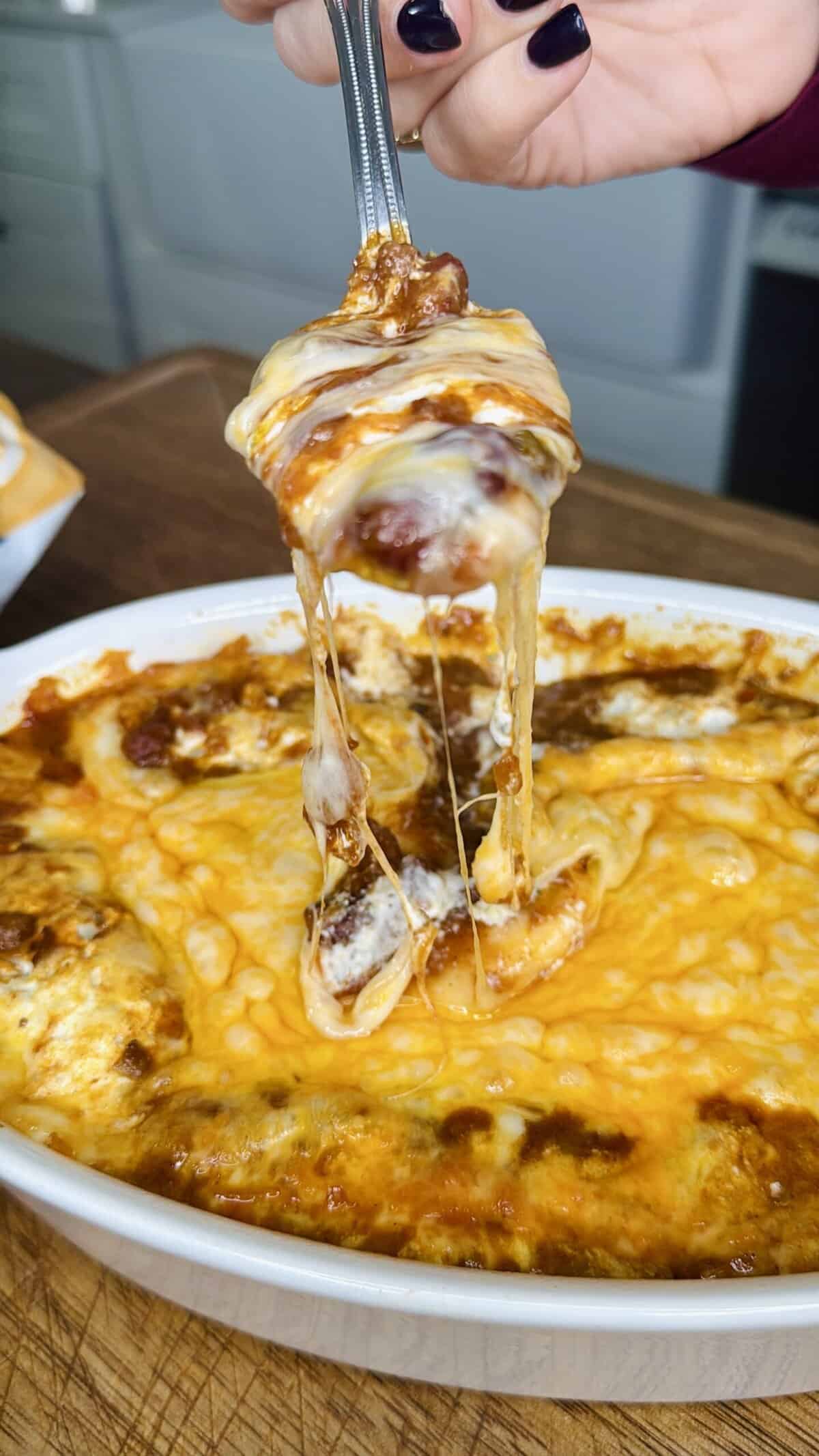 A hand lifts a cheesy, saucy serving from a casserole dish filled with golden-brown baked pasta—one of My Top 10 Christmas Recipes for Christmas Eve—with melted cheese stretching from the dish.