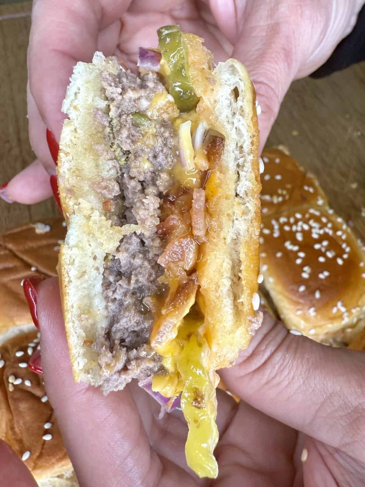 A close-up of hands holding a half-eaten cheeseburger with visible layers of beef patty, melted cheese, pickles, onions, tomato, lettuce, and sauce in a sesame seed bun—perfect inspiration for My Top 10 Christmas Recipes for Christmas Eve.