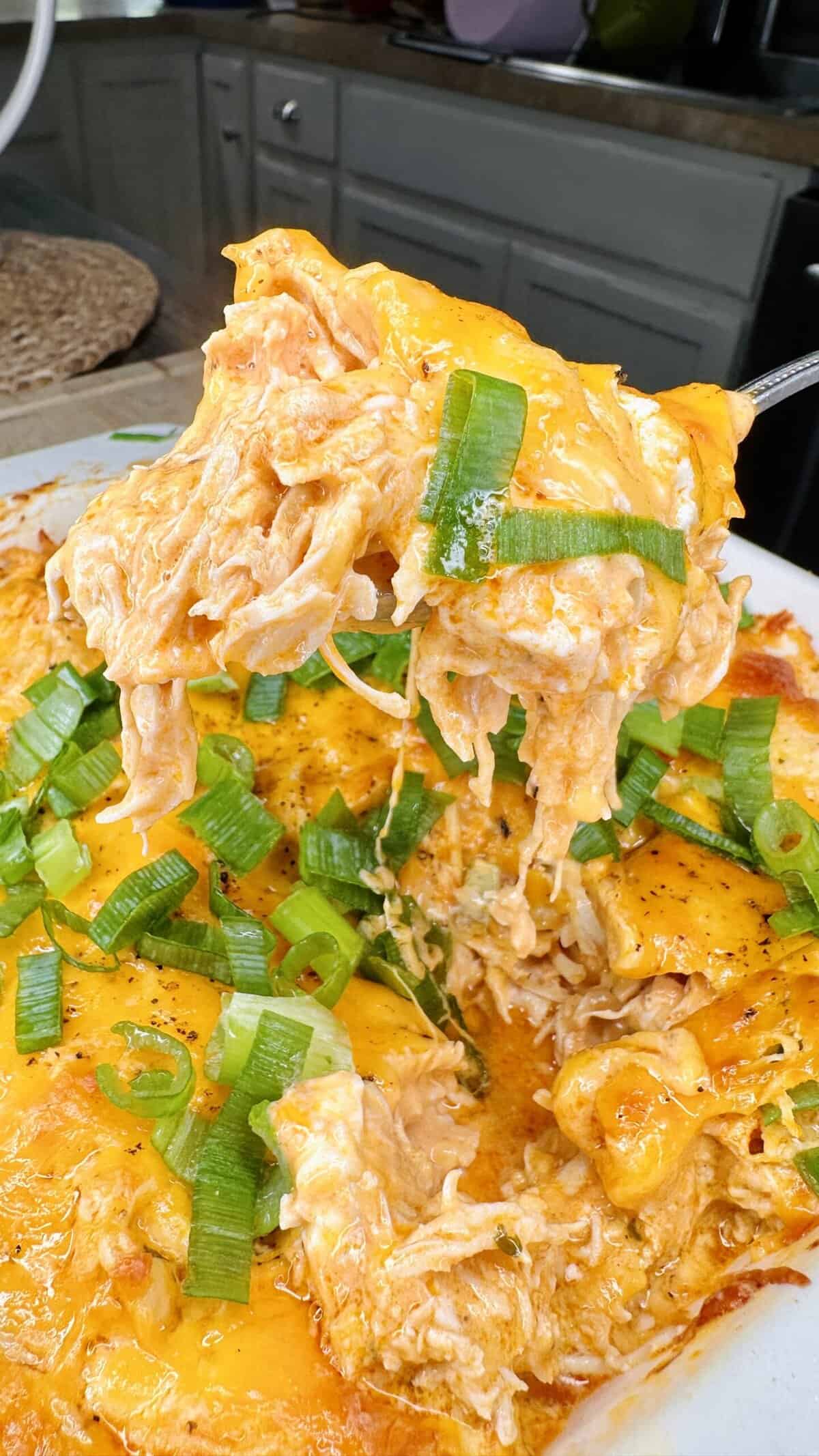 A close-up of a cheesy buffalo chicken casserole, one of My Top 10 Christmas Recipes for Christmas Eve, topped with sliced green onions. A serving is lifted out, revealing shredded chicken, melted cheese, and creamy orange buffalo sauce.