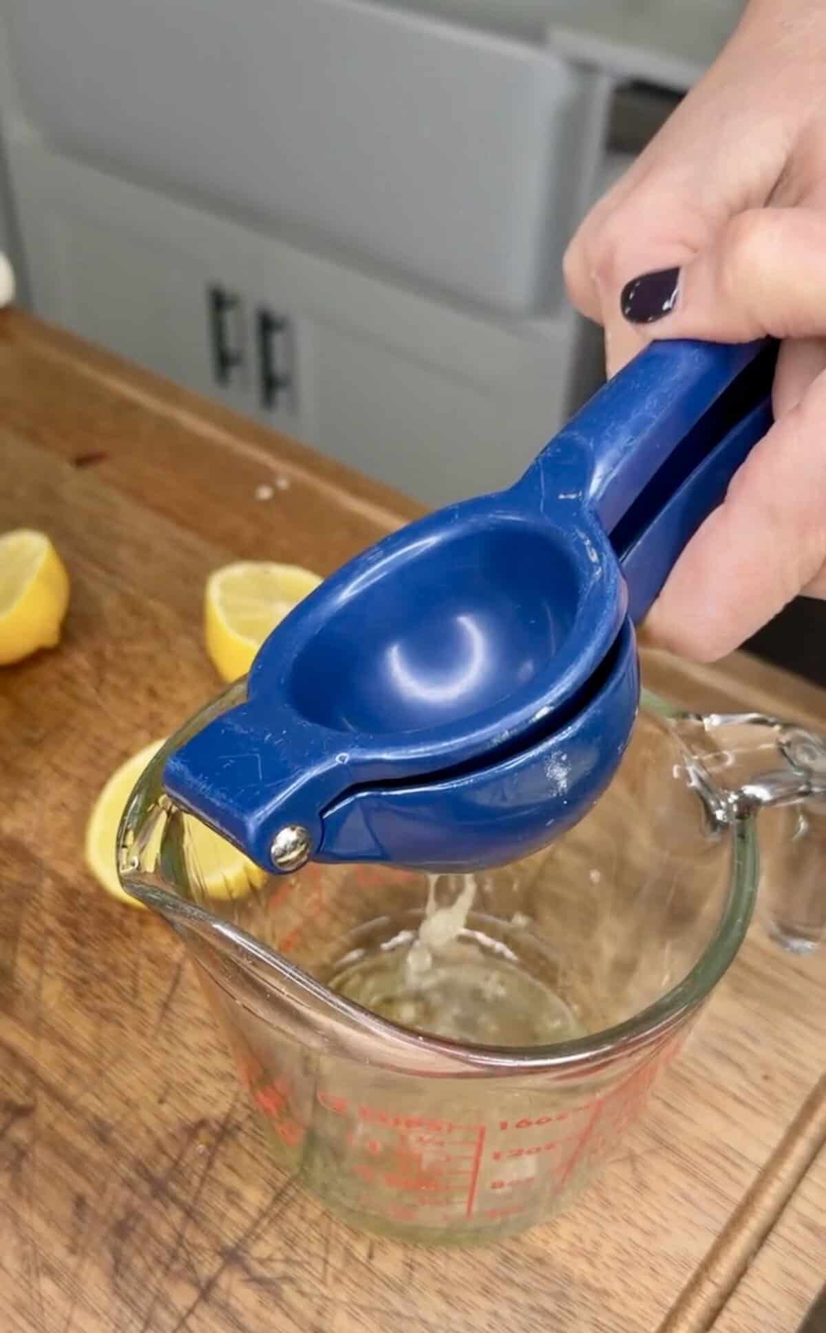 A person squeezes a blue lemon squeezer over a glass measuring cup on a wooden countertop, prepping fresh juice for Greek Restaurant-Style Lemon Roasted Potatoes with halved lemons in the background.
