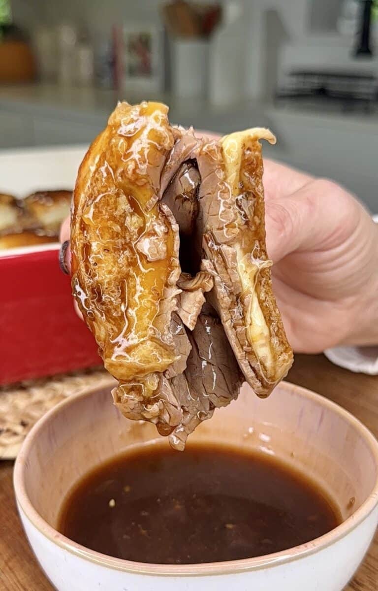 A hand holds a sandwich filled with melted cheese and sliced beef, dipped into brown au jus sauce—a cozy kitchen scene perfect for My Top 10 Christmas Recipes for Christmas Eve.