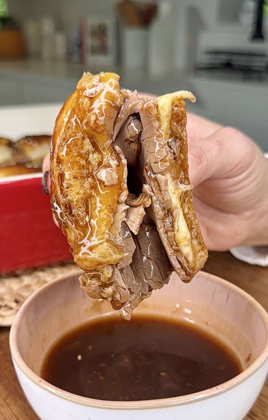 A hand holds a sandwich filled with melted cheese and sliced beef, dipped into brown au jus sauce—a cozy kitchen scene perfect for My Top 10 Christmas Recipes for Christmas Eve.