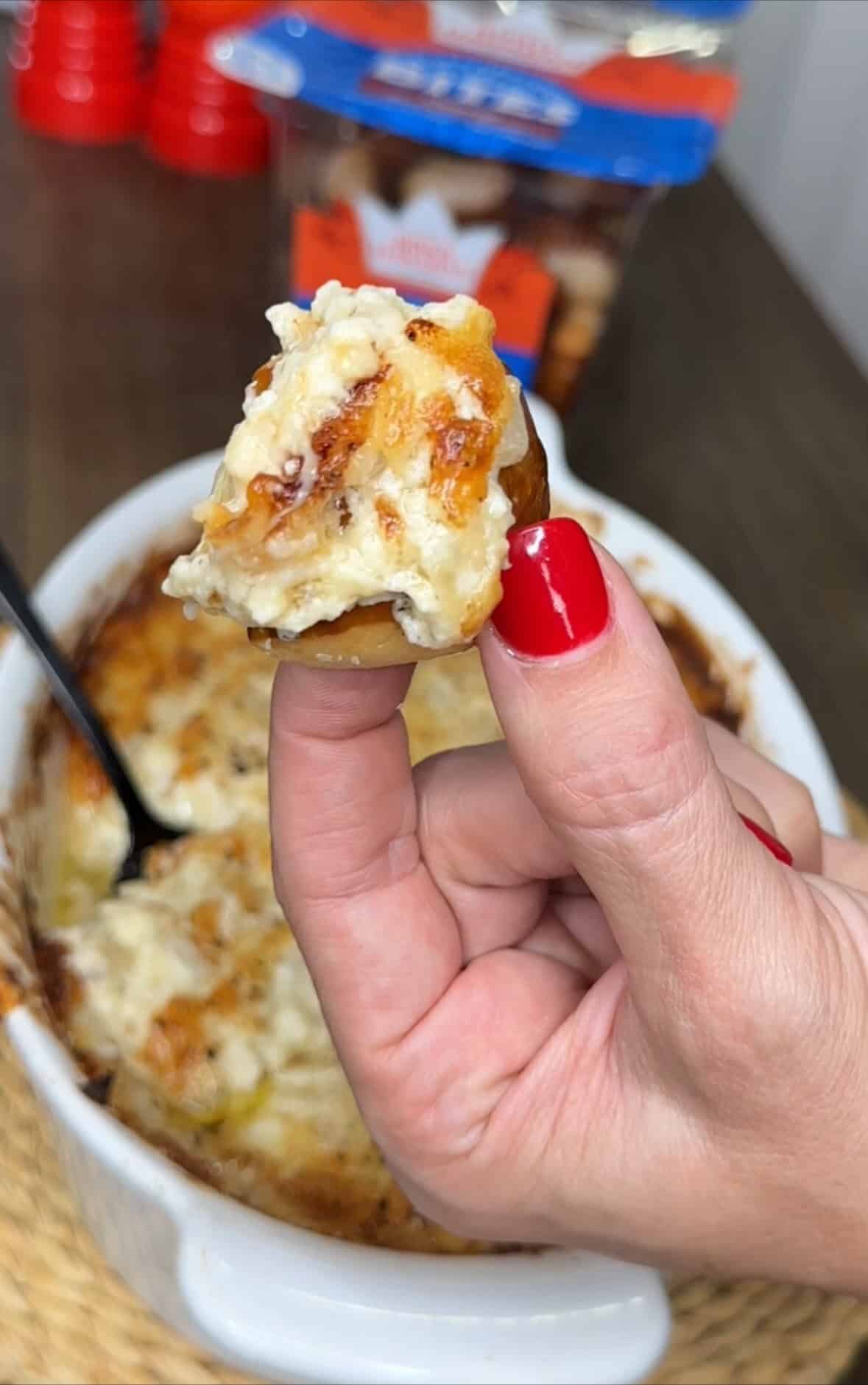 A hand with red nail polish holds a piece of bread topped with creamy, cheesy dip—one of My Top 10 Christmas Recipes for Christmas Eve—with a dish of baked dip and a spoon in the background.