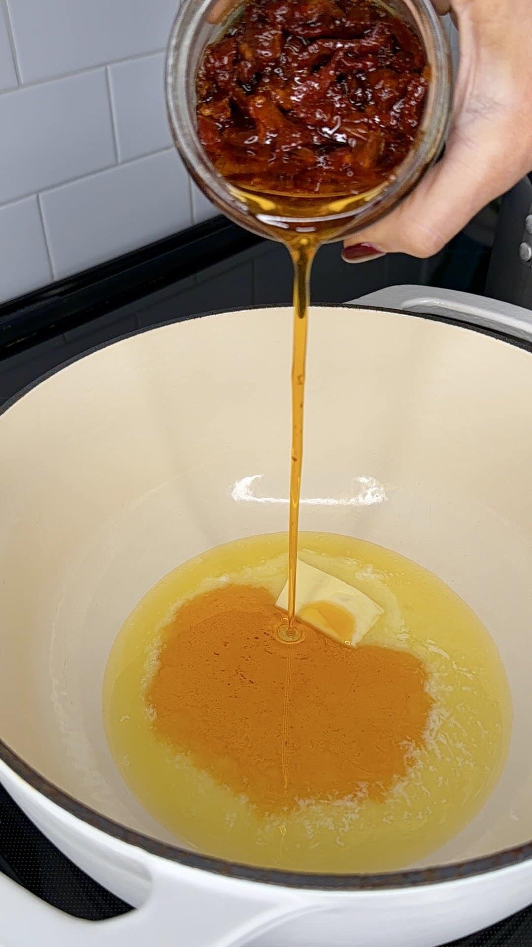 A hand pours chili oil with bits of chili from a glass jar into a white pot with melted butter on a stovetop, against a backdrop of white tiled wall.