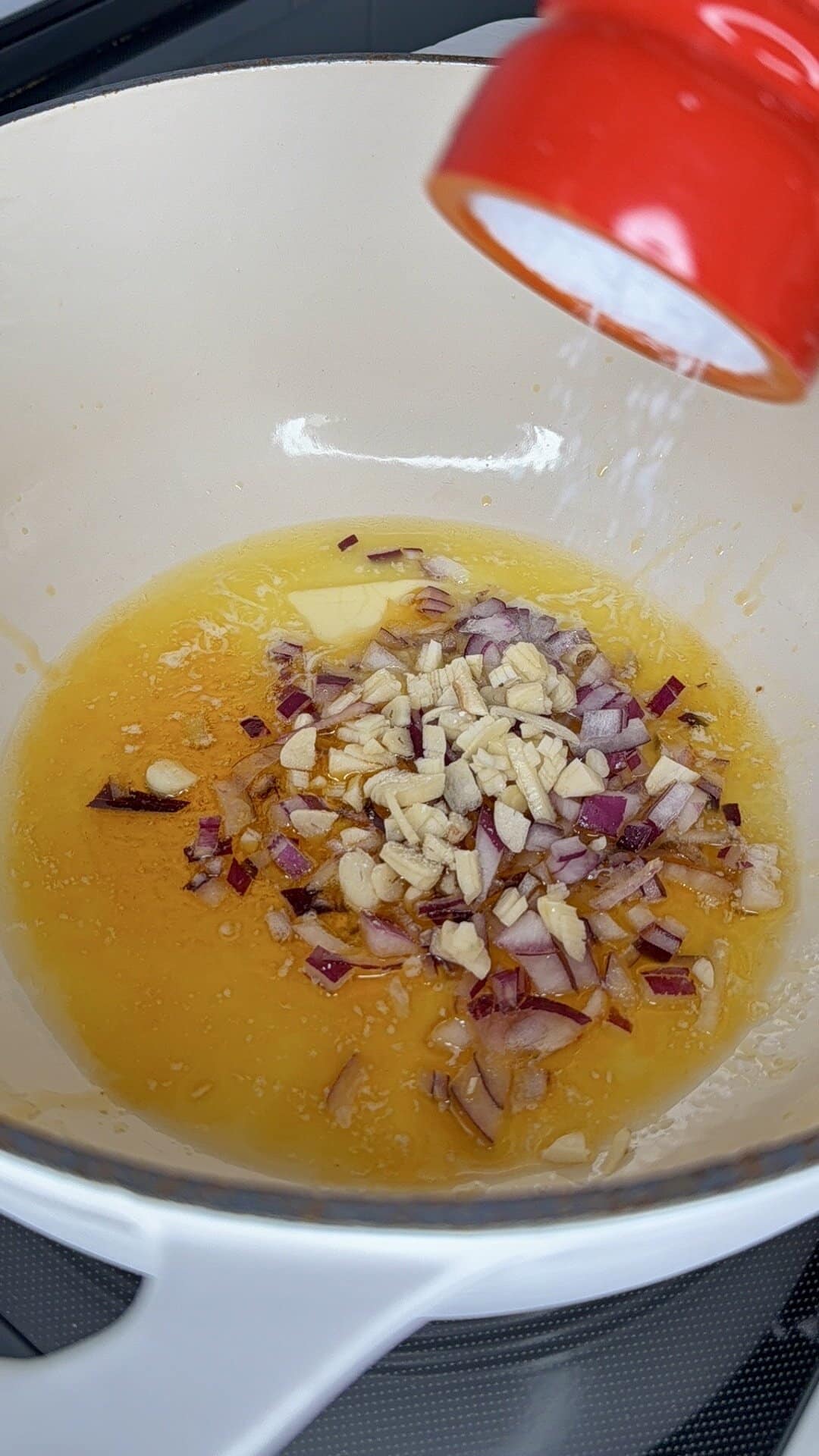 Chopped red onion and garlic cooking in melted butter in a white pot, with salt being sprinkled in from a red container.