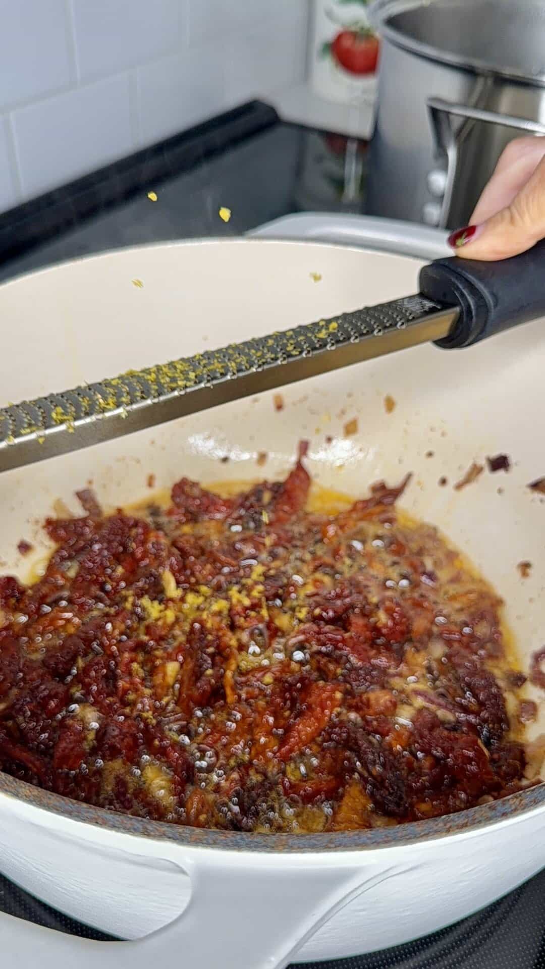 A hand grates lemon zest into a white pot with simmering tomato sauce on a stove. A stainless steel pot and a tomato-themed container are visible in the background.