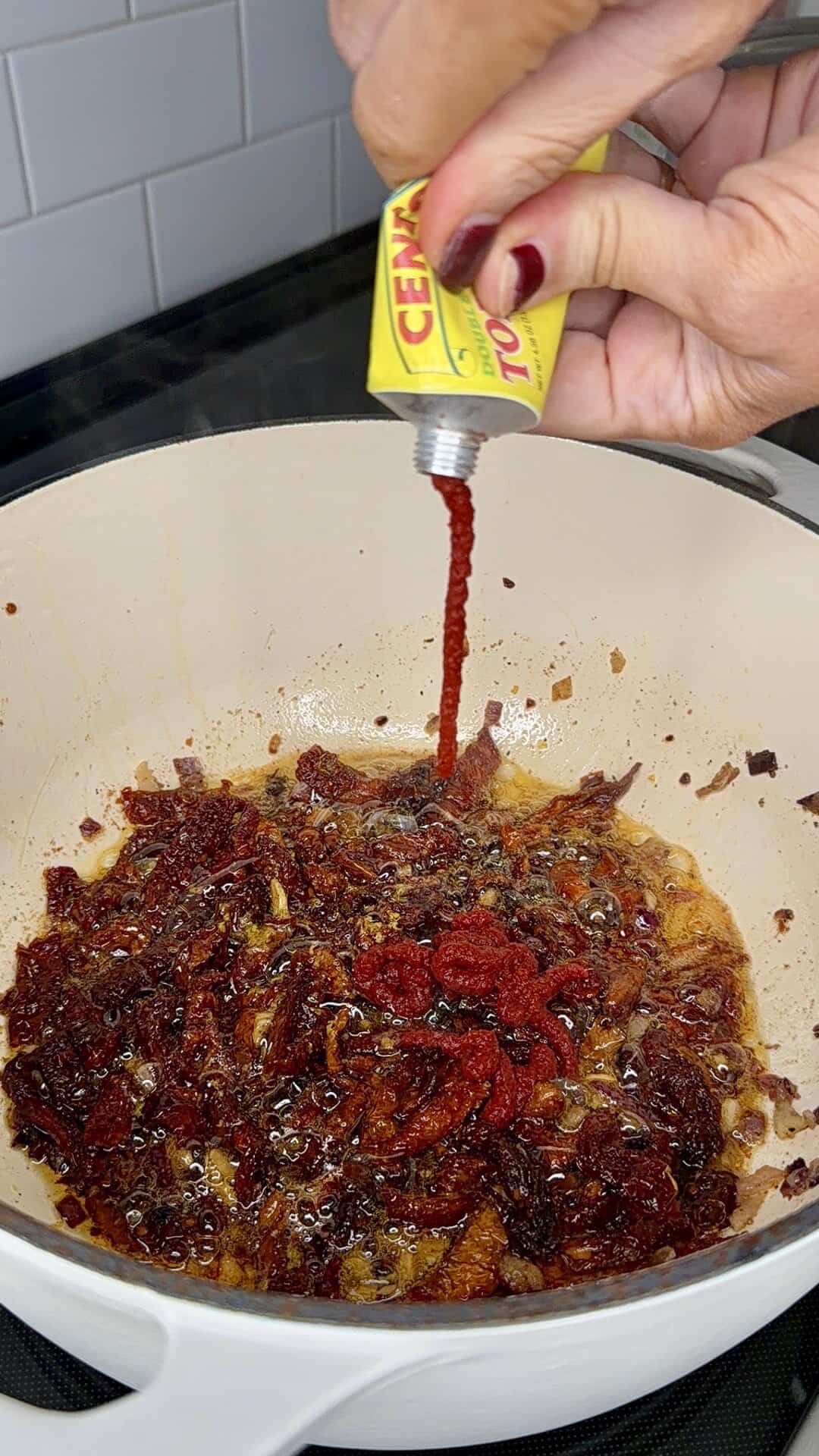 A hand squeezes tomato paste from a yellow tube into a pot with sizzling onions, garlic, and dried chilies on a stovetop.