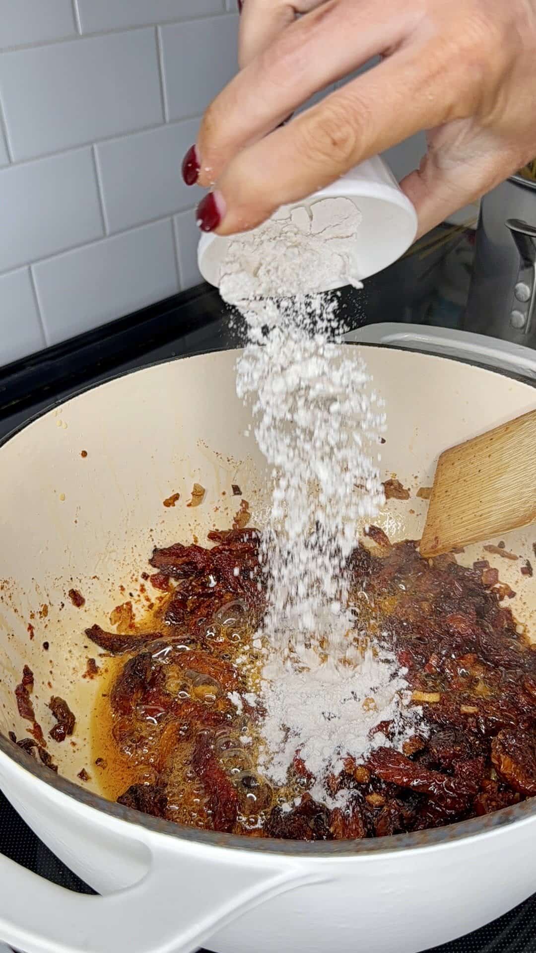 A hand pours flour from a small white container into a cream-colored pot with sautéed vegetables and spices, while stirring with a wooden spoon on a stovetop.