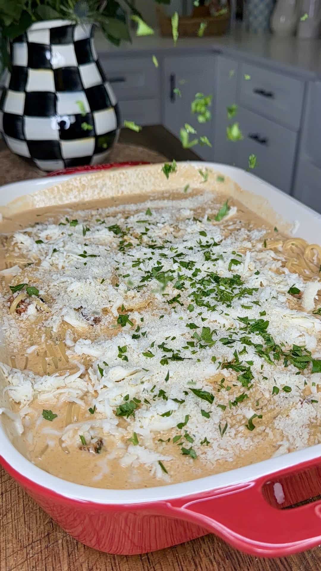 A red baking dish filled with creamy marry me baked spaghetti, topped with grated cheese and freshly chopped herbs, sits on a kitchen counter. Parsley is being sprinkled from above. A black-and-white checkered vase is in the background.