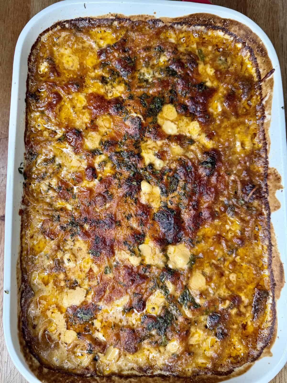 A close-up view of a baked casserole in a white rectangular dish, featuring the golden-brown, slightly crispy top of marry me baked spaghetti with melted cheese, herbs, and savory sauce visible on the surface.