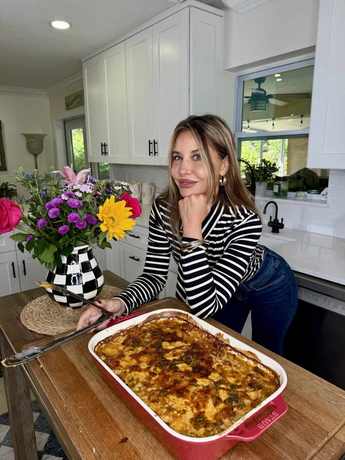 A woman in a striped shirt smiles and leans on a kitchen counter next to a red dish of marry me baked spaghetti. A colorful bouquet of flowers sits nearby, with white cabinets and a window in the background.