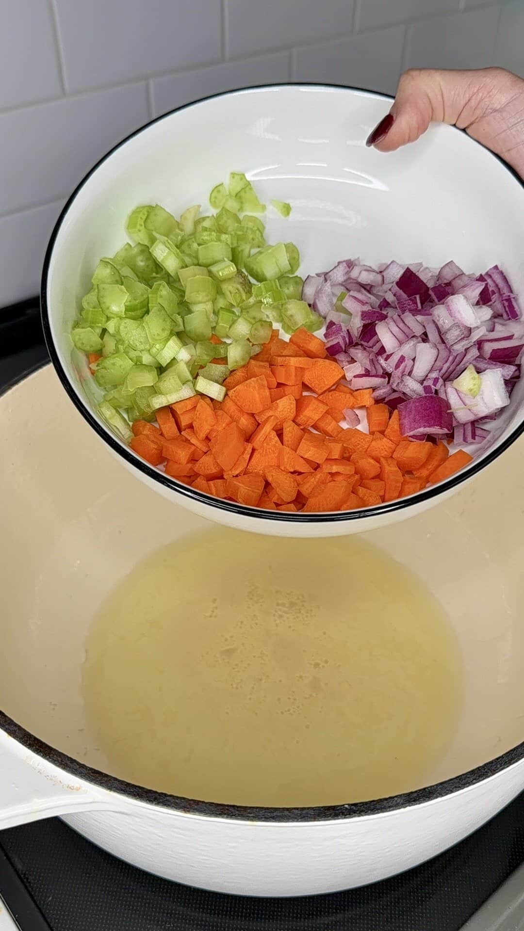 A hand holds a bowl filled with chopped celery, carrots, and red onion over a white pot on a stove, ready to add the vegetables to the heated oil inside the pot.