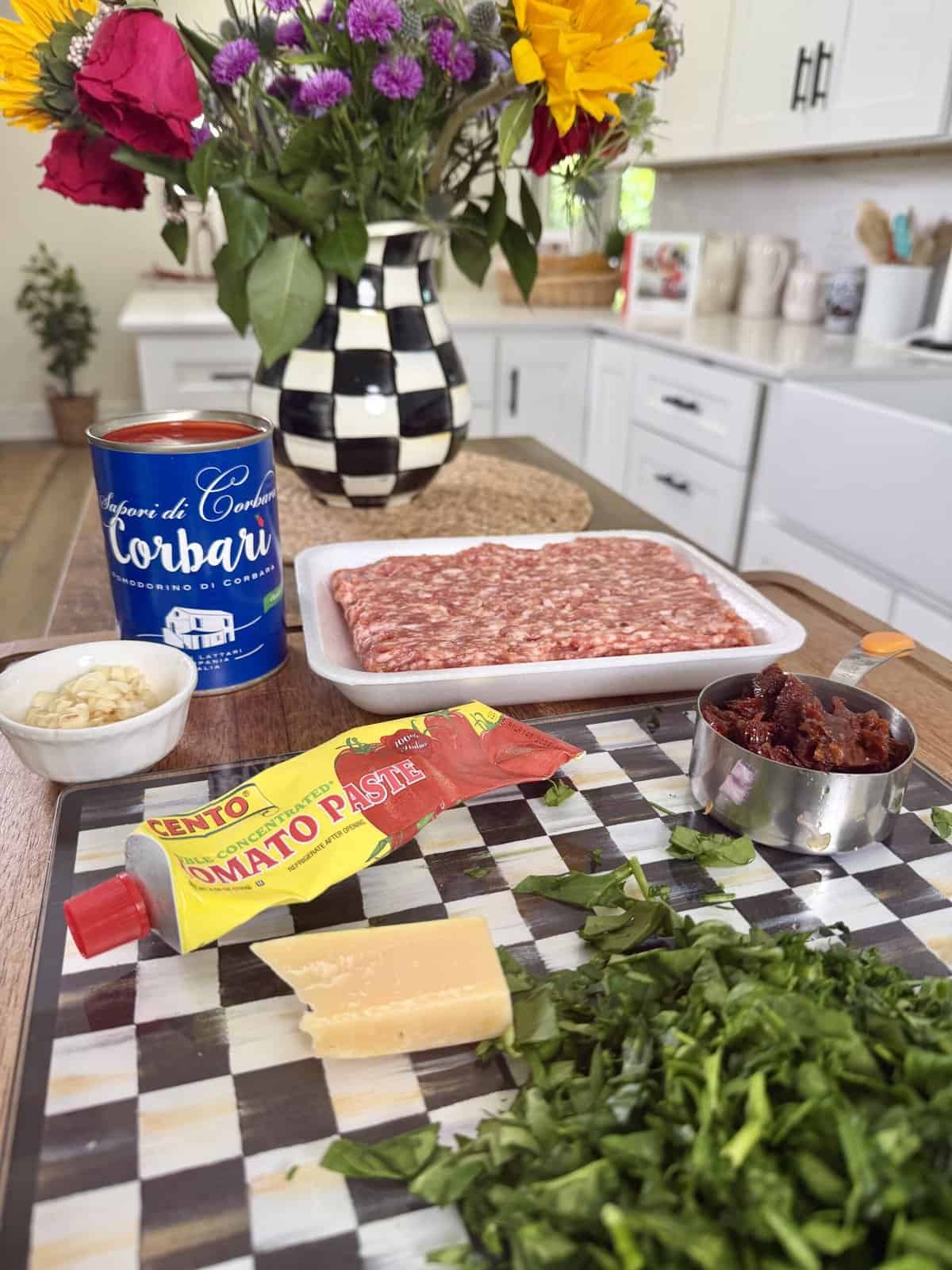 A kitchen countertop displays Italian sausage, tomato paste, canned tomatoes, chopped greens, grated Parmesan, minced garlic, and sundried tomatoes. A vase of colorful flowers sits in the background.