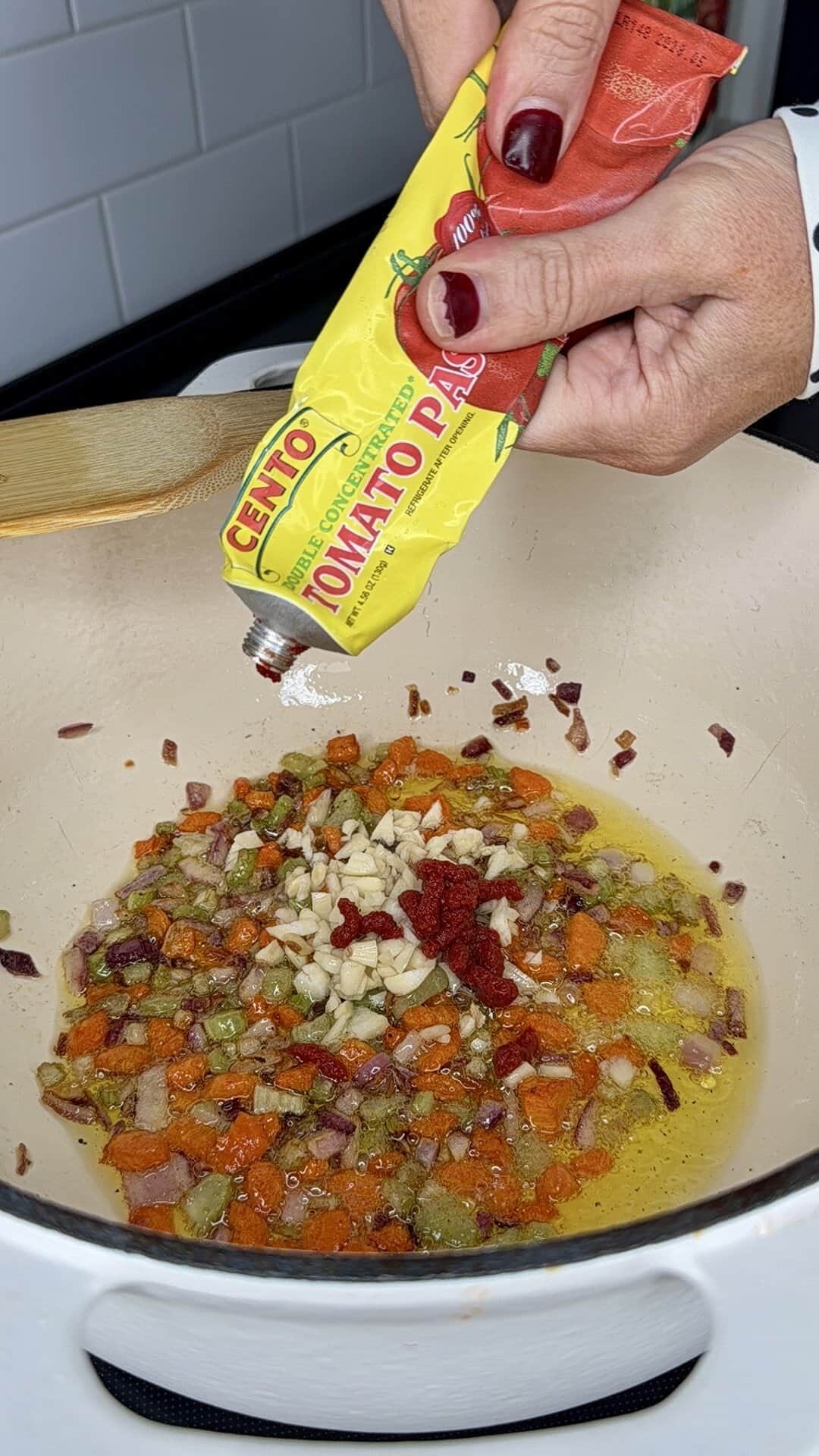A hand squeezes tomato paste from a yellow tube into a pot with sautéed diced onions, carrots, celery, garlic, and oil on a stovetop. A wooden spoon rests on the edge of the pot.
