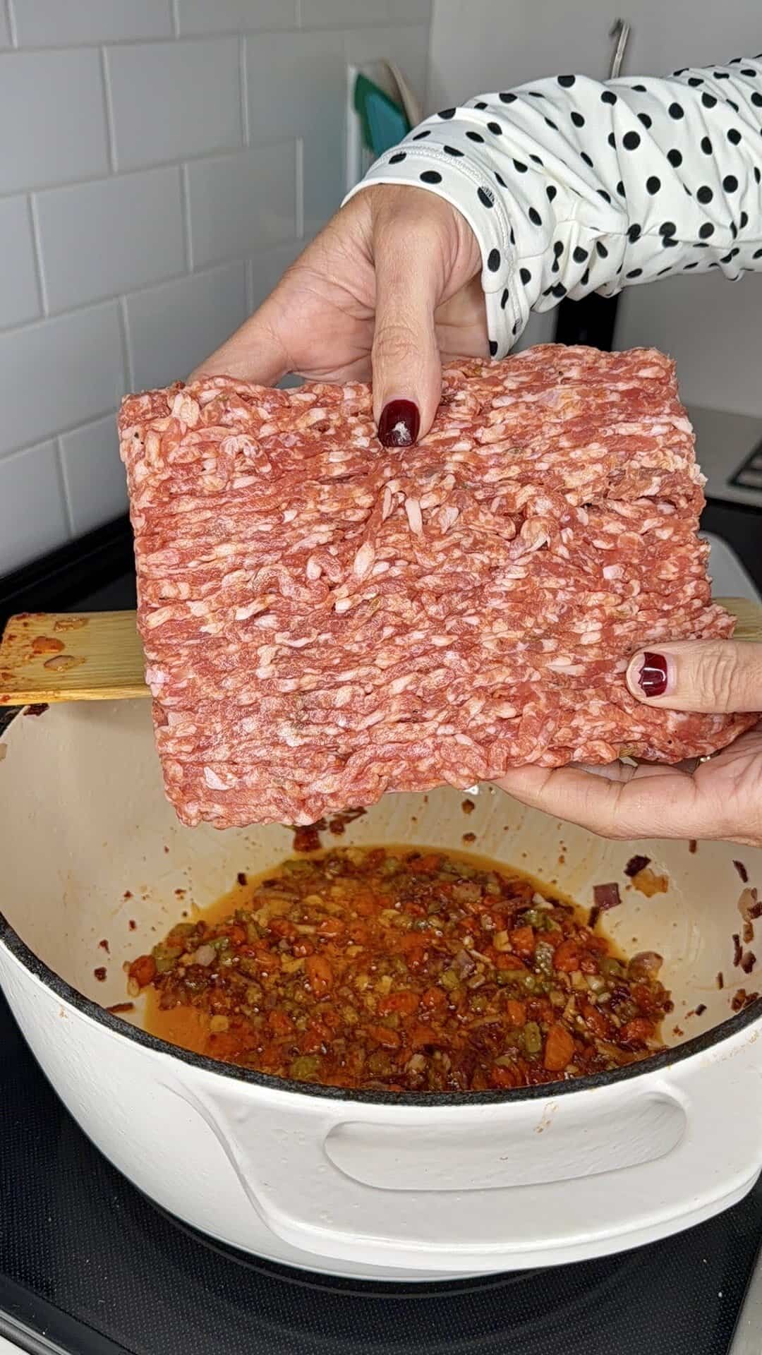 A person holding a block of raw ground meat over a white pot filled with cooked vegetables and sauce on a stovetop. The persons nails are painted dark red, and they are wearing a white shirt with black polka dots.