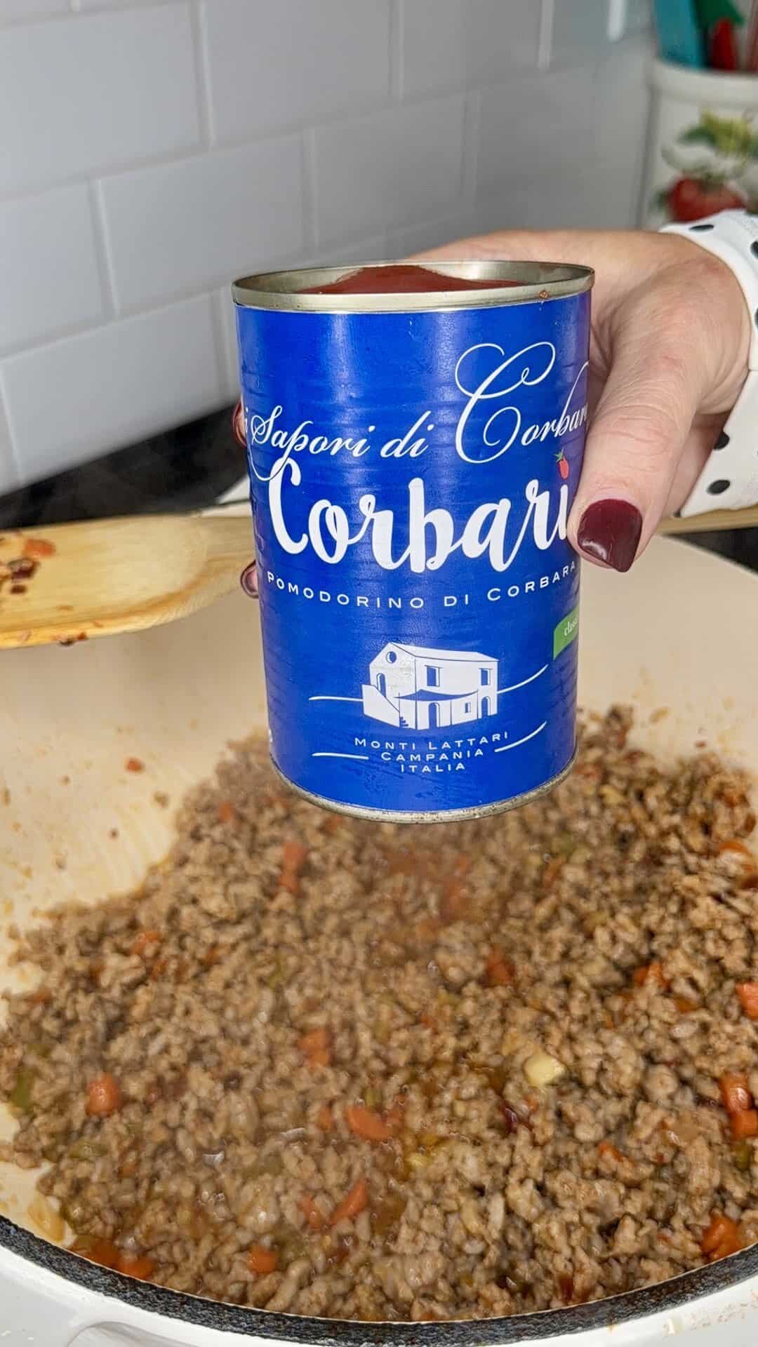 A hand with red nails holds a blue can of Corbara tomatoes above a pot of ground meat and vegetables cooking on a stove, with a wooden spoon stirring the mixture.