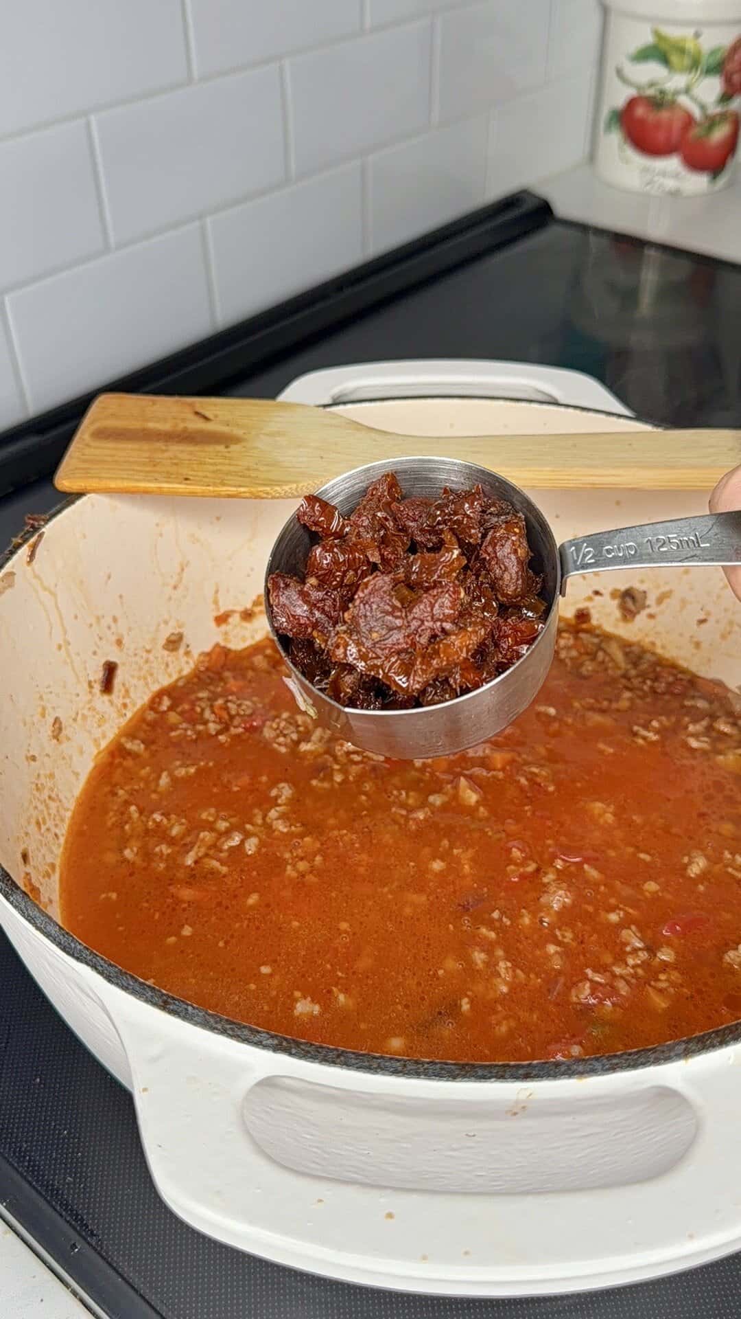 A hand holds a metal measuring cup filled with chopped sun-dried tomatoes over a white pot of simmering tomato and ground meat sauce on a stovetop. A wooden spoon rests on the pot.