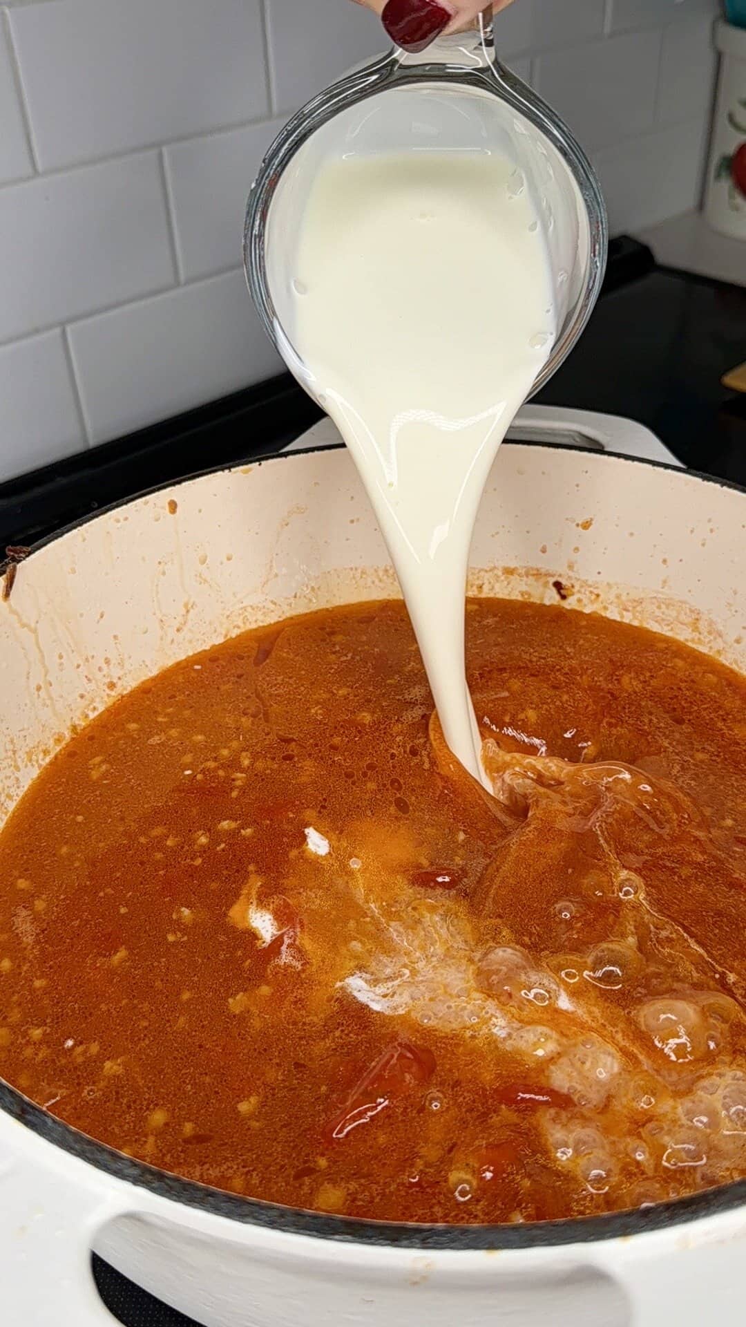 A hand pours cream from a glass container into a pot of red tomato-based soup on a stovetop, with white subway tile in the background.