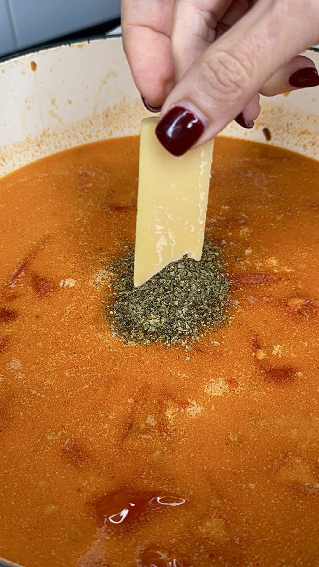 A hand with red nail polish holds a piece of Parmesan cheese above a pot of tomato soup, sprinkling dried herbs into the simmering mixture.
