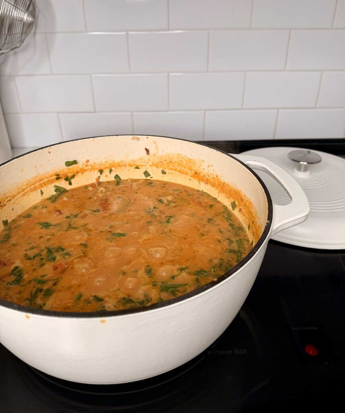 A large white Dutch oven sits on a stovetop, filled with a bubbling orange-red stew or soup with visible greens and herbs. The white lid rests nearby, and the background shows a white tiled backsplash.