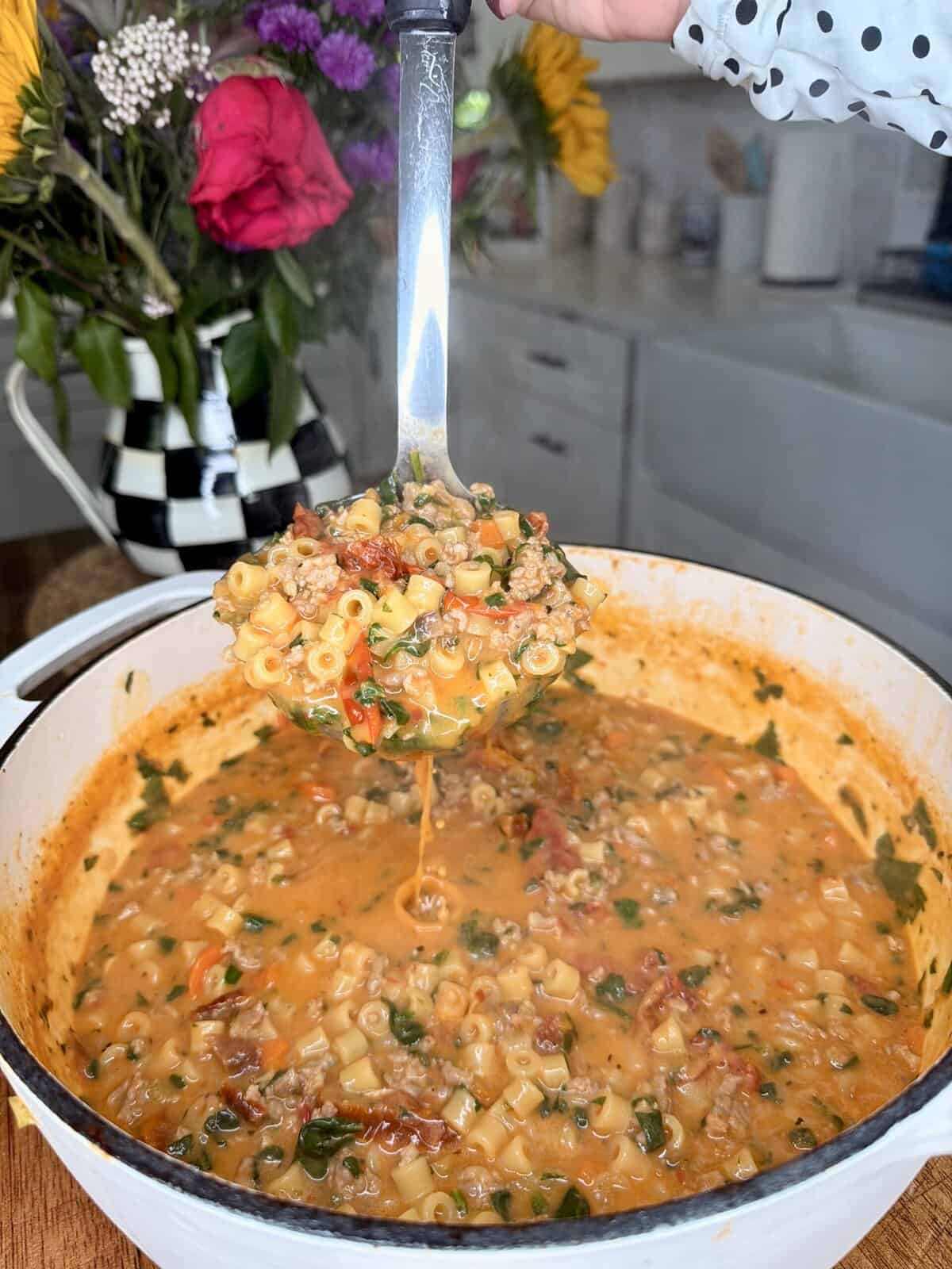 A ladle lifts a serving of hearty pasta soup with ground meat, small pasta, spinach, and tomatoes from a white pot in a bright kitchen. A vase of colorful flowers sits in the background.