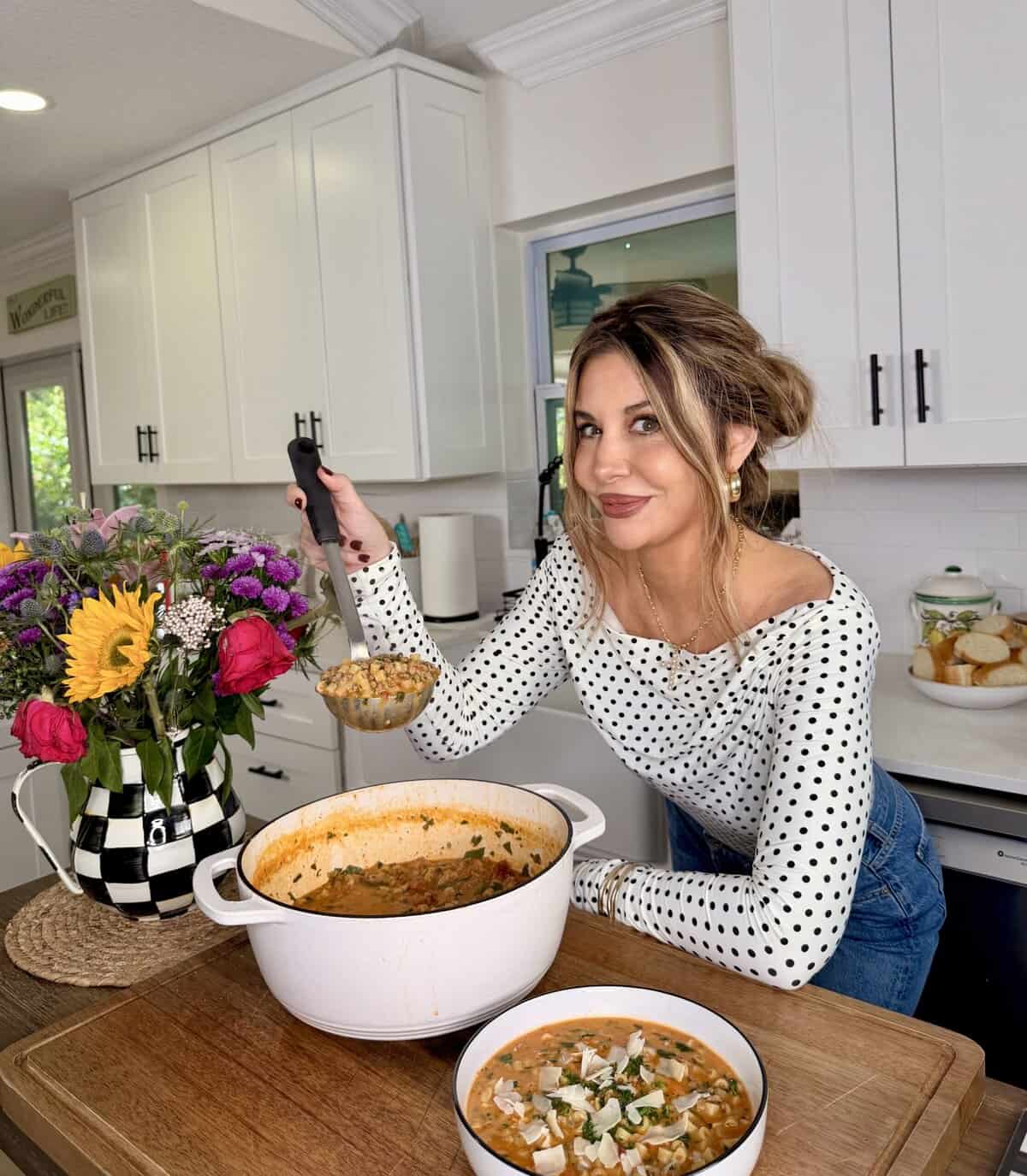 A woman in a white polka dot top serves soup from a large white pot in a bright kitchen. A bowl of soup and a colorful bouquet of flowers are on the wooden counter beside her.