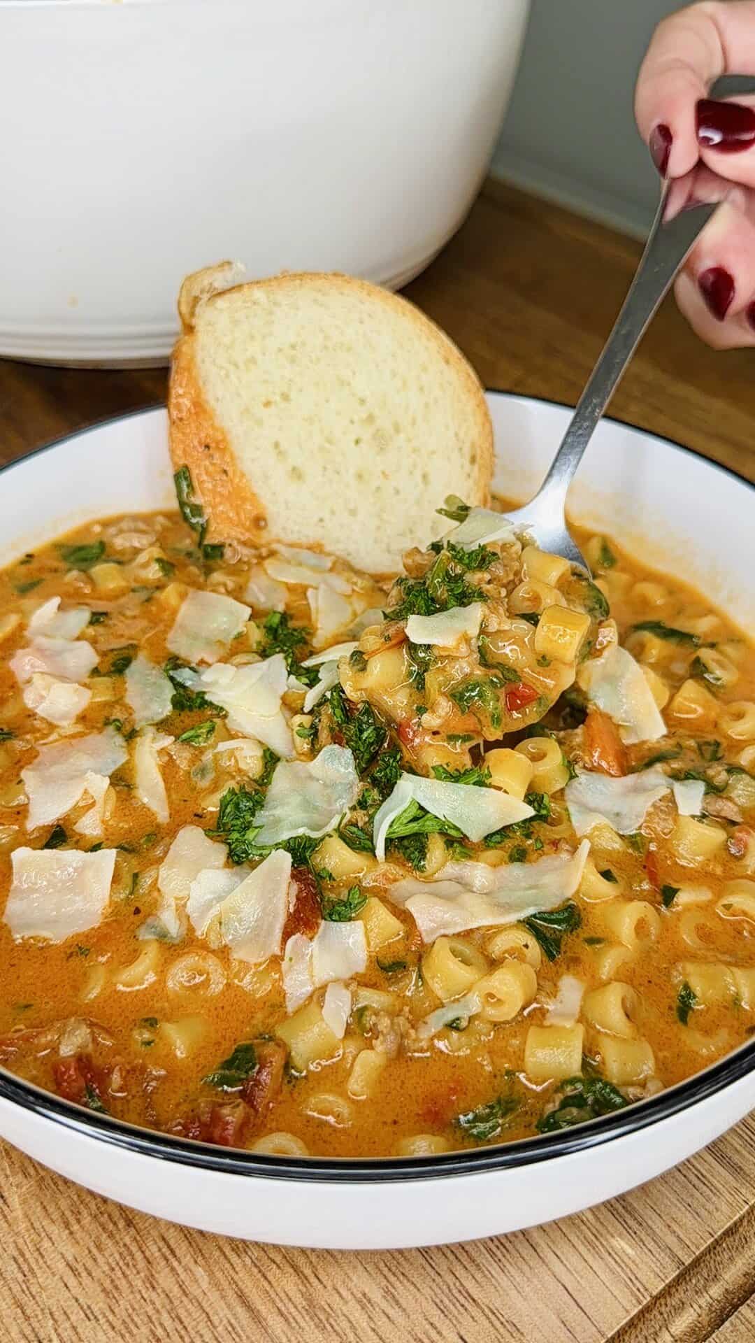 A bowl of hearty soup with pasta, ground meat, greens, and tomato broth, topped with shaved parmesan. A hand holds a spoonful above the bowl, and a slice of bread rests on the bowls rim.