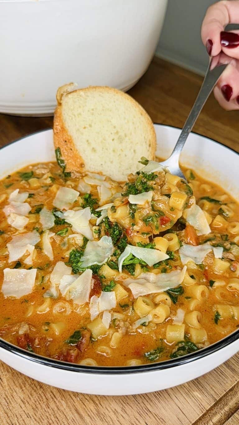 A bowl of creamy pasta soup topped with shaved cheese and leafy greens, served with a slice of bread. A hand is holding a spoonful of the soup above the bowl.