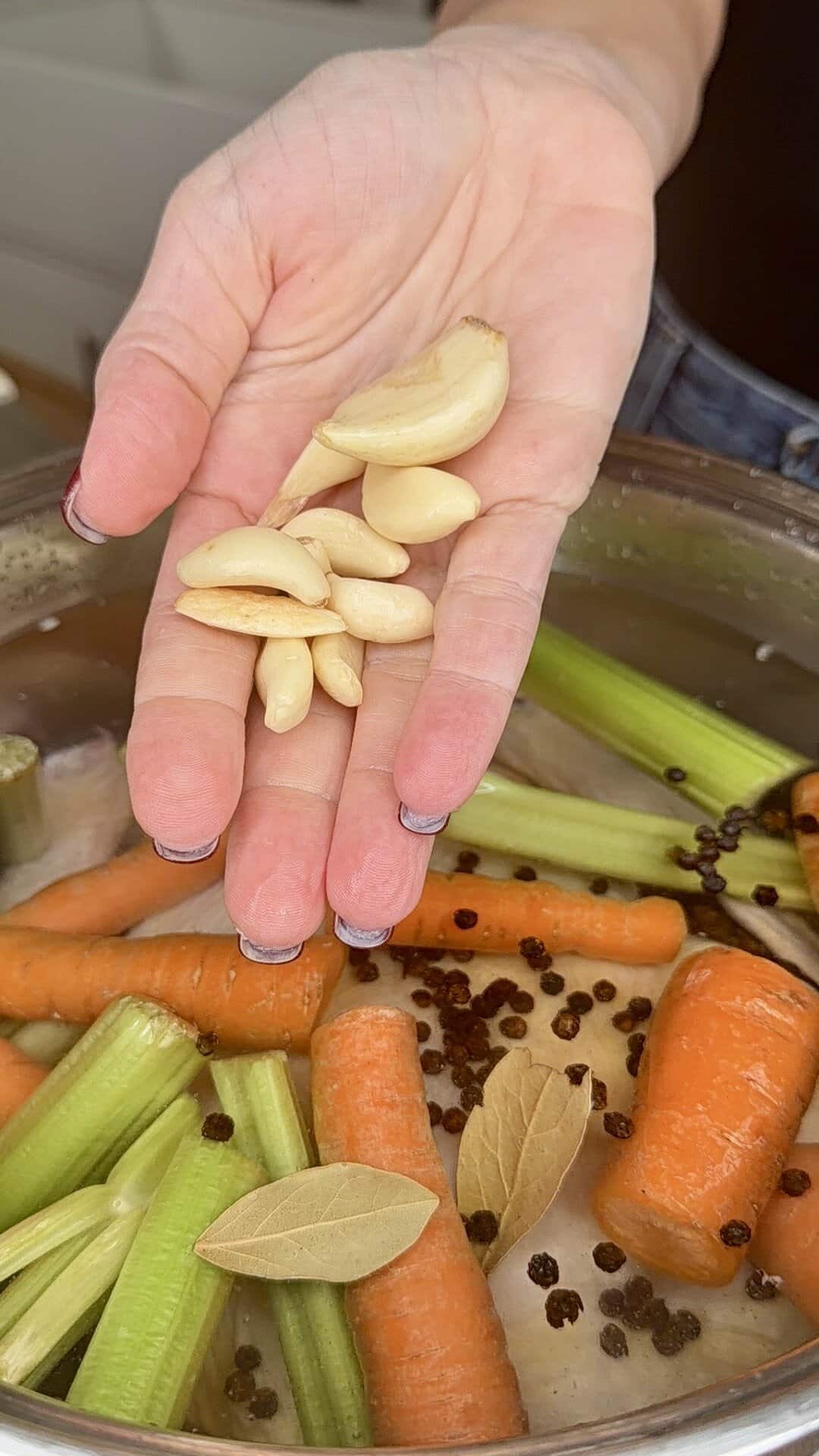 A hand holding peeled garlic cloves over a pot filled with carrots, celery, bay leaves, and peppercorns, preparing ingredients for a homemade broth or soup.
