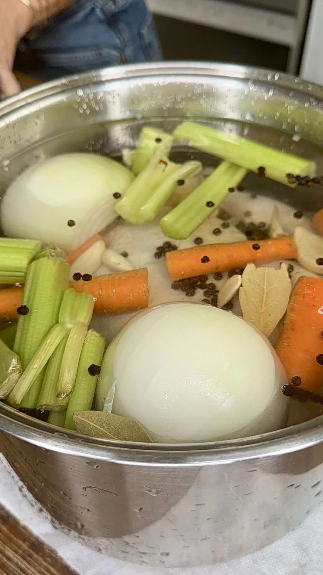 A stainless steel pot filled with water, halved onions, chopped carrots and celery, bay leaves, garlic cloves, and whole black peppercorns, likely being prepared for making broth or stock.