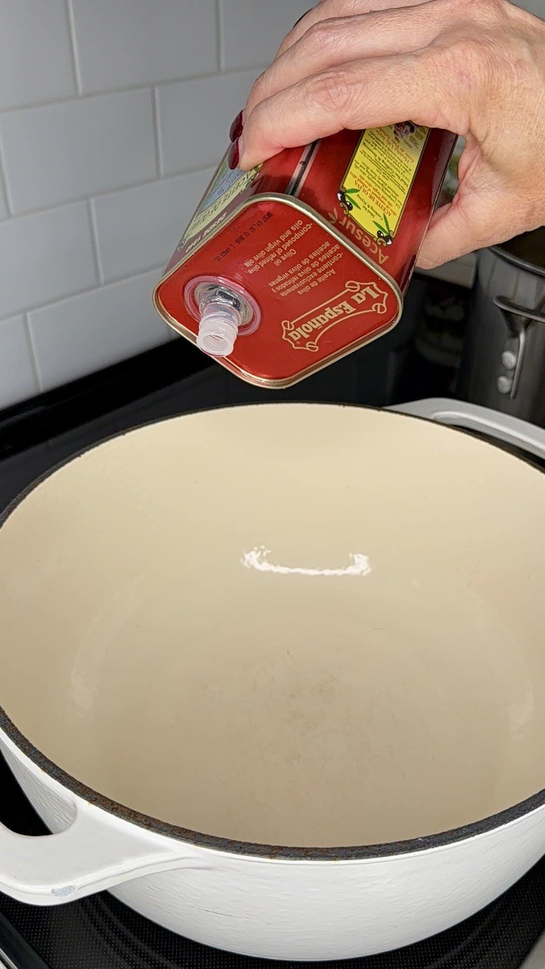 A hand pours olive oil from a red can into a large white Dutch oven on a stovetop, with white tiled backsplash in the background.
