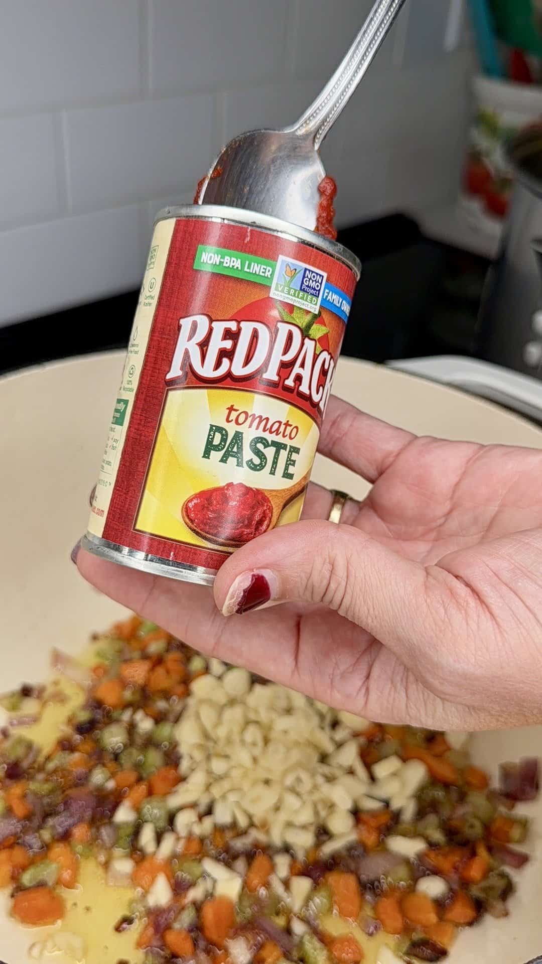 A hand holds a can of Red Pack tomato paste with a spoon, above a pot containing chopped vegetables sautéing in oil on a stove.