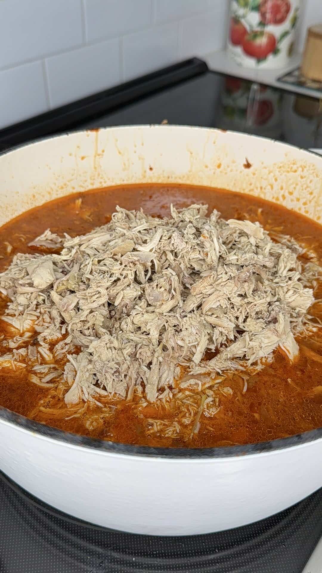 Shredded meat piled in the center of a pot filled with red, seasoned broth, sitting on a stove. Kitchen items, including a container with a tomato design, are visible in the background.