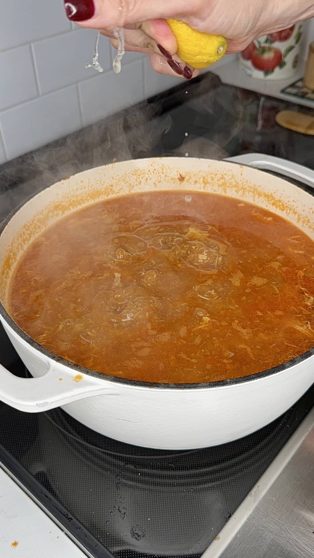 A hand squeezes a lemon over a steaming pot of red soup or stew in a white Dutch oven on a stovetop.