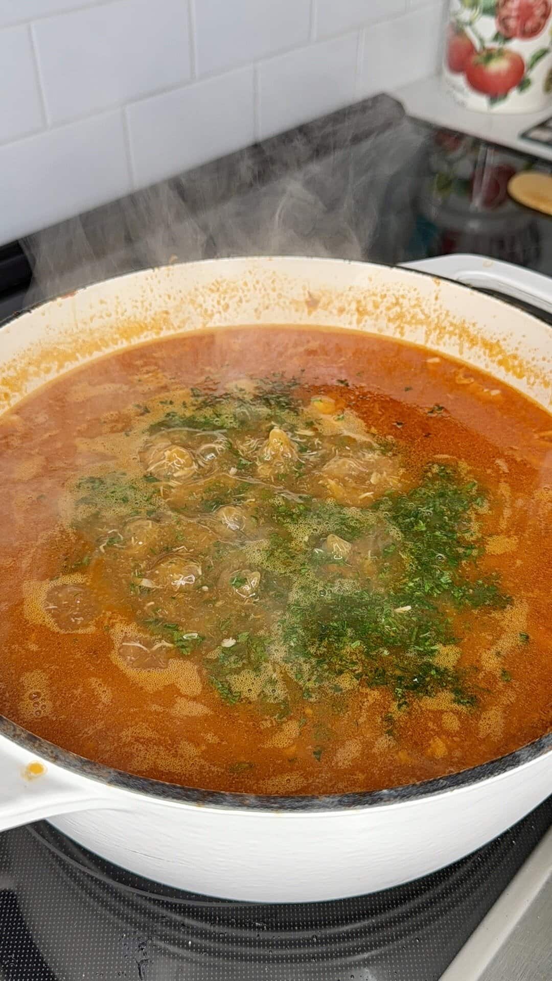 A white pot filled with bubbling red soup garnished with chopped herbs, simmering on a stovetop with steam rising. White subway tiles and kitchen items are visible in the background.