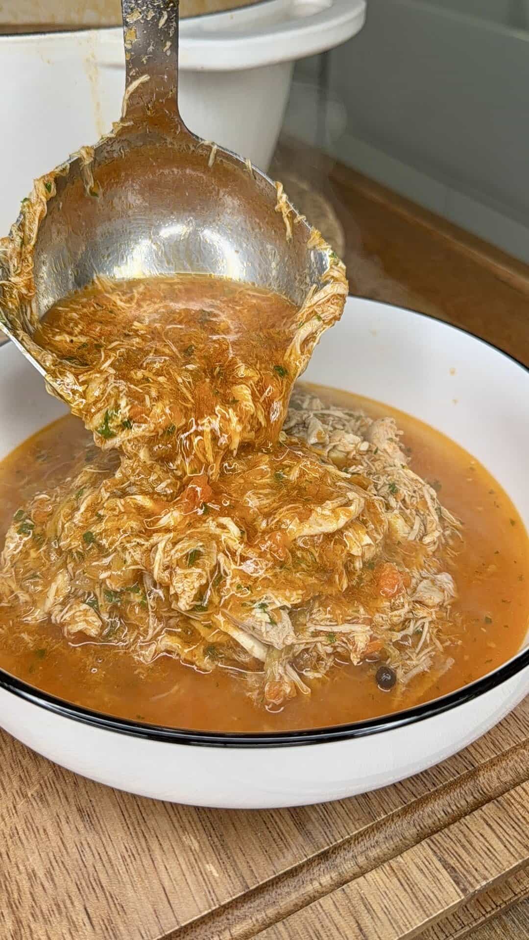 A ladle pours shredded meat in a red, spiced sauce into a white bowl with a black rim, set on a wooden surface. Steam rises from the hot dish, suggesting it is freshly cooked.