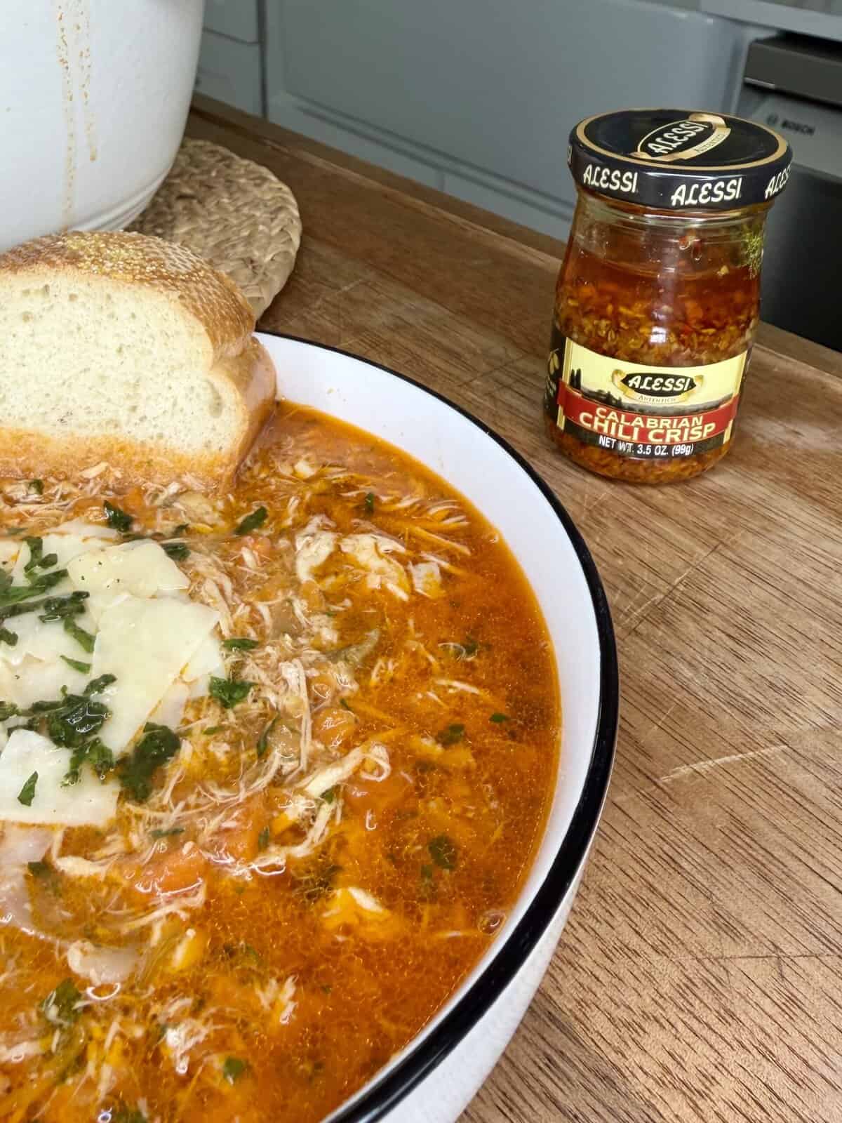 A bowl of shredded chicken soup topped with herbs and cheese sits on a wooden counter with a slice of bread. In the background, there is a jar of Acessi Calabrian chili peppers.