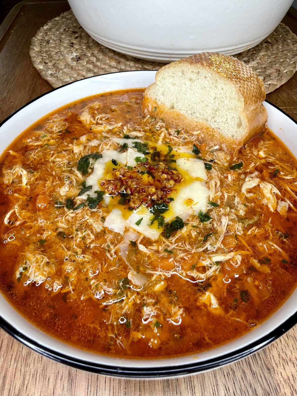 A bowl of spicy red chicken soup garnished with herbs, shredded cheese, and chili oil, served with a slice of crusty bread resting on the edge of the bowl.