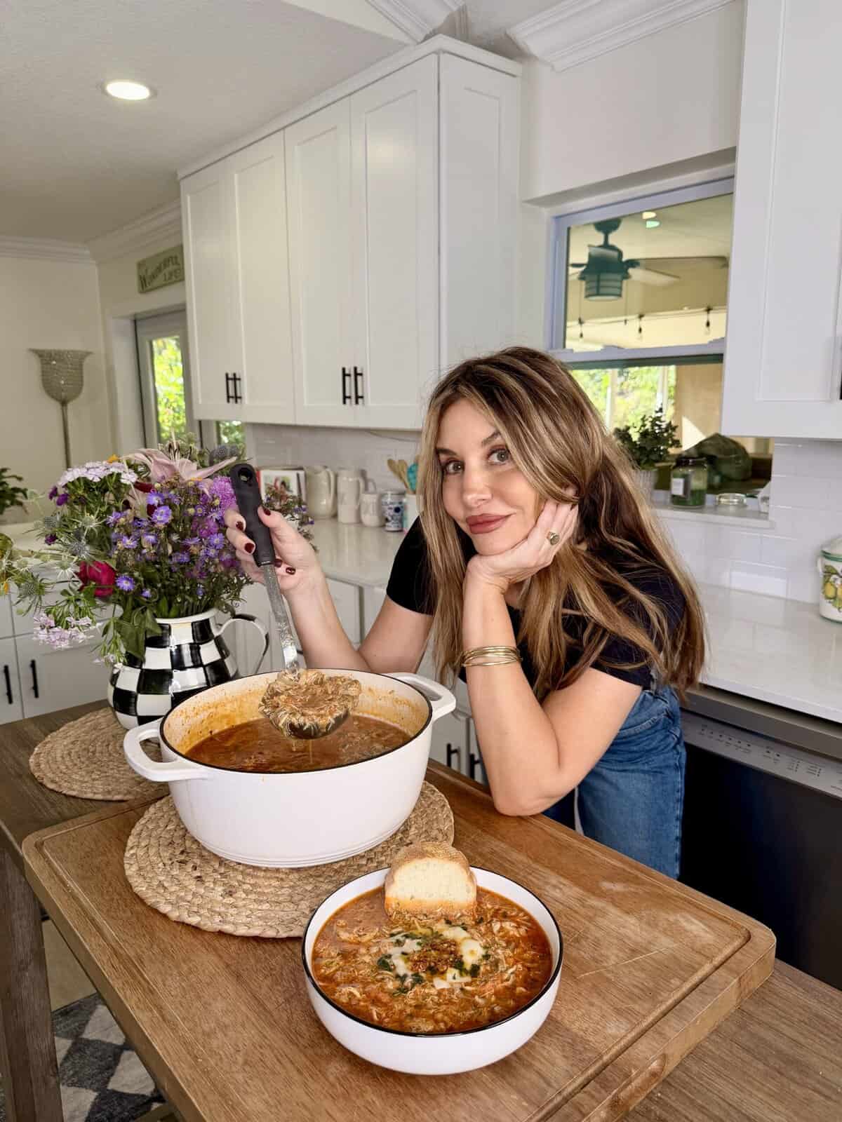 A woman with long brown hair, wearing a black shirt and jeans, smiles while serving soup from a white pot in a bright kitchen. A bowl of soup with bread sits in front of her on the counter.