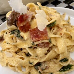 A close-up of Creamy Bacon Mushroom Fettuccine with crispy bacon pieces, wilted spinach, sautéed mushrooms, and shaved Parmesan cheese on a white plate.