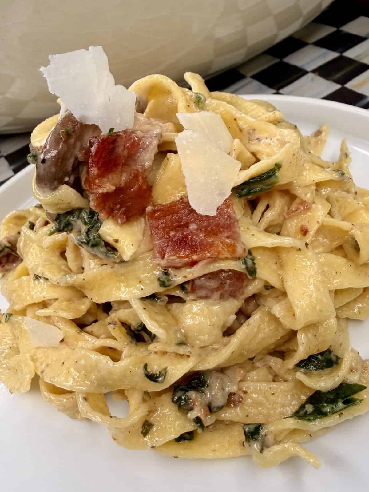 A close-up of Creamy Bacon Mushroom Fettuccine with crispy bacon pieces, wilted spinach, sautéed mushrooms, and shaved Parmesan cheese on a white plate.