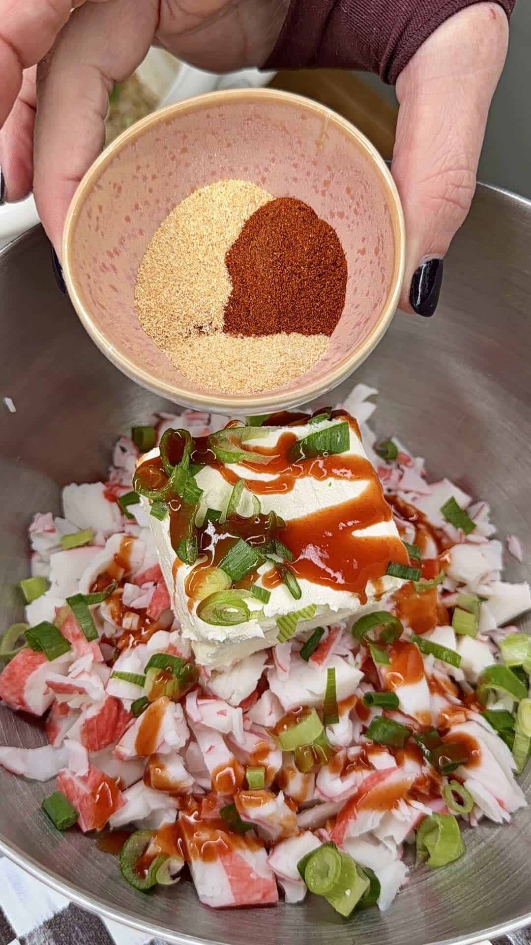 A hand holds a small bowl with powdered spices above a metal mixing bowl containing chopped imitation crab, cream cheese, sliced green onions, and hot sauce.