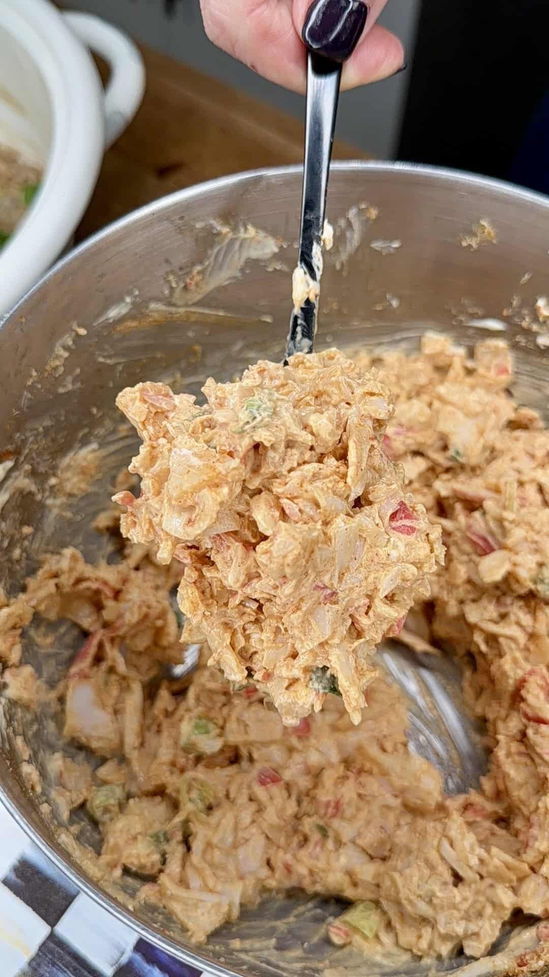 A close-up of a metal mixing bowl with a creamy, chunky dip or salad mixture being lifted by a spoon. The mixture includes visible pieces of vegetables and has a reddish, orange hue.